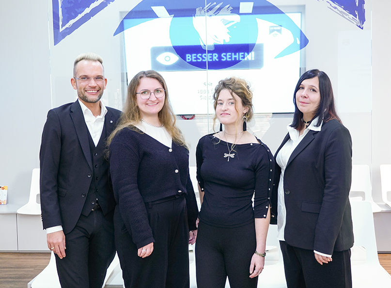Augenoptiker-Team (bestehend aus Optikchef und drei Mitarbeiterinnen) vor Glaswand mit Logo sowie Bildschirm