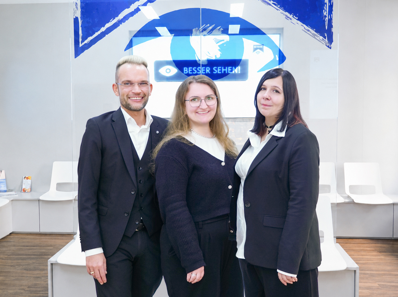 Optikerteam bestehend aus Optikermeister mit Brille und zwei Linsenspezialistinnen vor Glaswand und Bildschirm