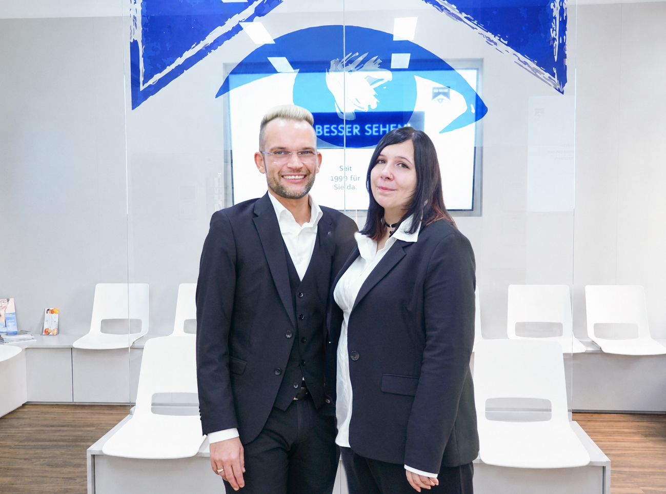Optikteam, bestehend auf Mann mit kurzen Haaren und Brille sowie Frau mit längeren Haaren vor Glaswand und Bilschirm