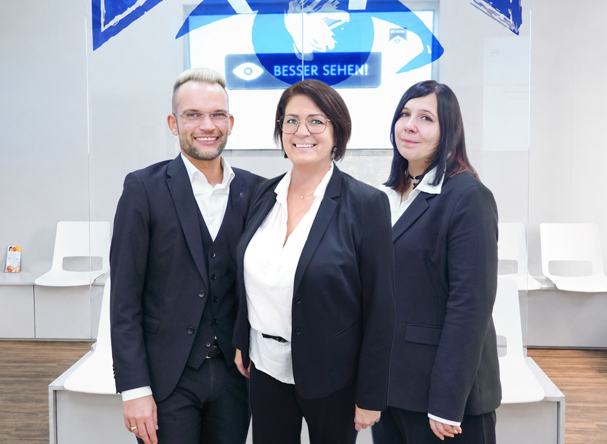 Optikerteam, bestehend aus einem Mann und zwei Frauen in Anzug, vor Warteraum mit Glaswand und Bildschirm